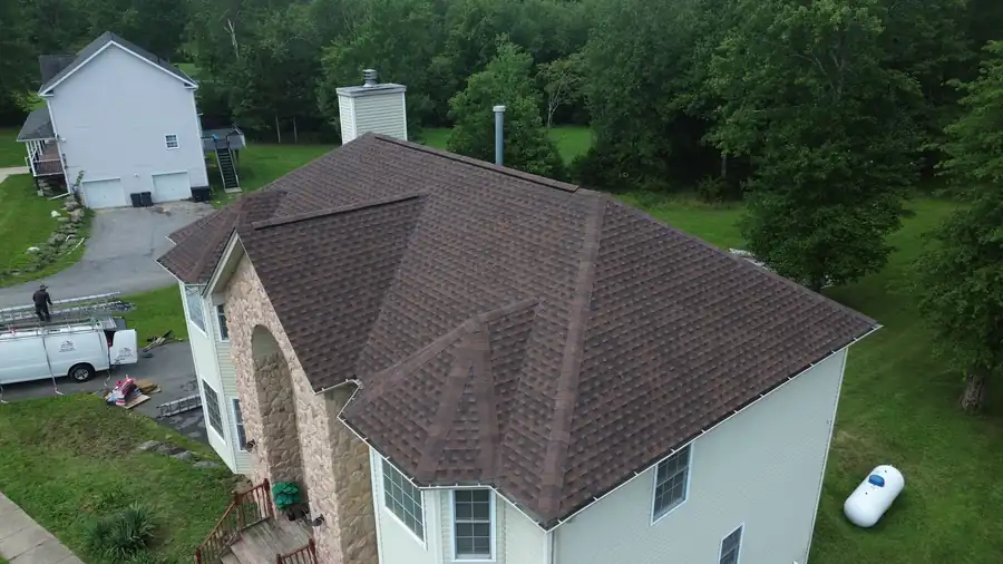 Drone view of a stone-front house under a new deep brown hip roof, woods behind